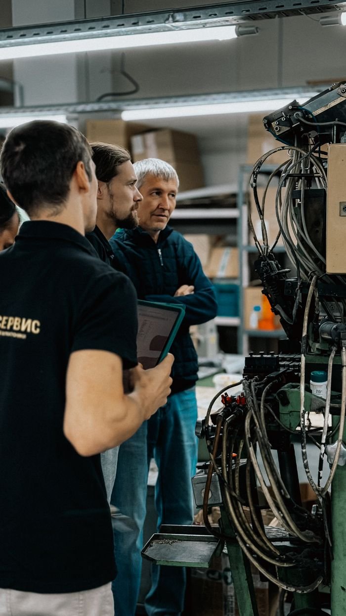 services-04 Three adults engaged in a discussion around complex machinery in a factory environment.